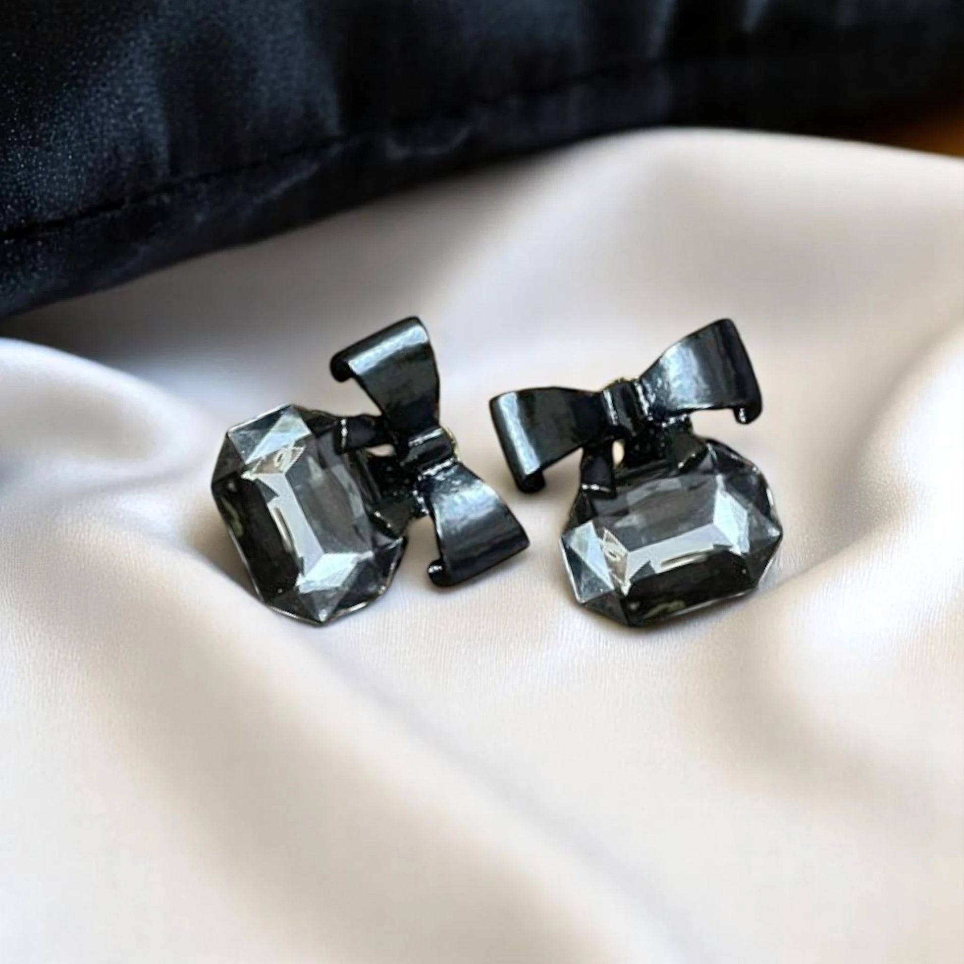 Elegant black bow earrings adorned with faceted gray gemstones, resting on a smooth white satin surface.