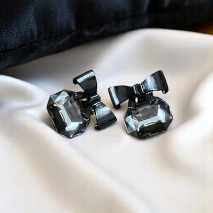 Elegant black bow earrings adorned with faceted gray gemstones, resting on a smooth white satin surface.