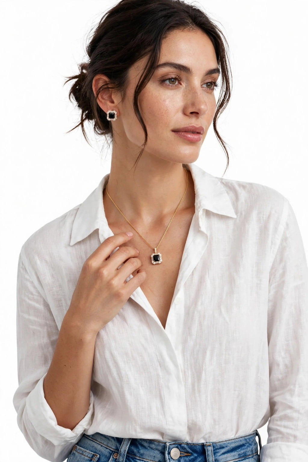 A woman wearing a delicate gold necklace with a square black and white stone pendant, matching stud earrings, and a white linen shirt against a plain white background.