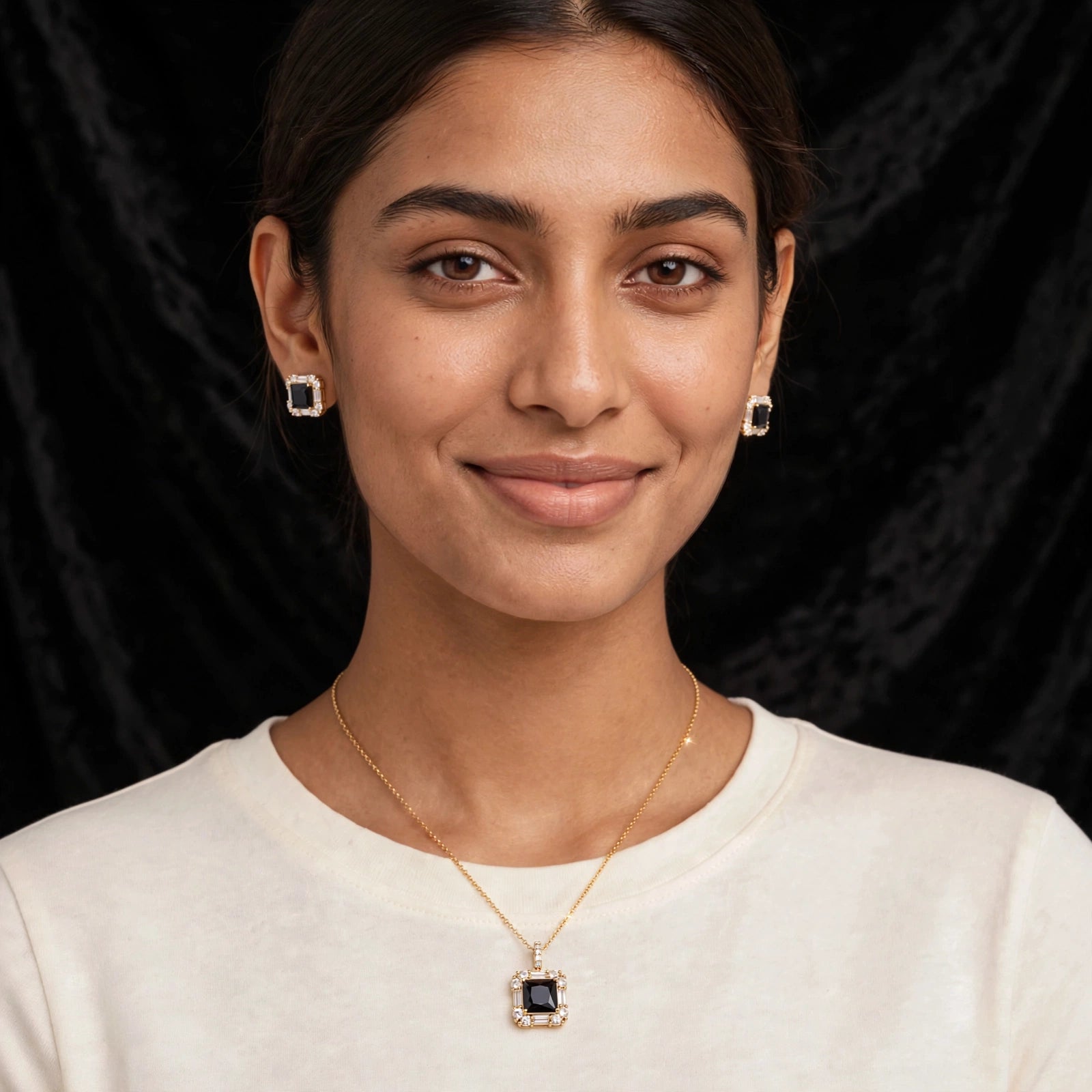 A woman wearing a gold necklace with a square black pendant and matching square stud earrings, shown against a dark textured background.
