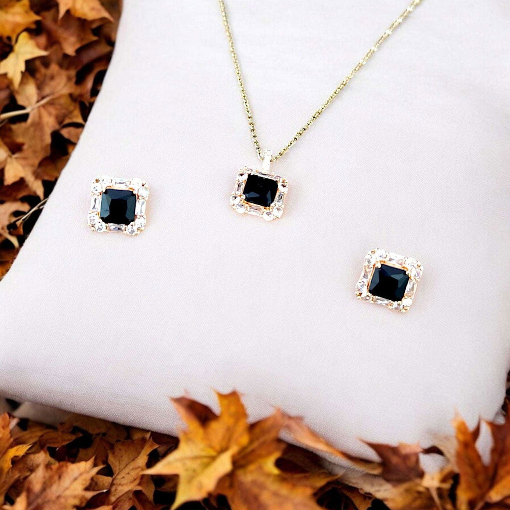 A gold necklace and matching stud earrings set with square black gemstones surrounded by small clear stones, displayed on a white cushion with autumn leaves around it.