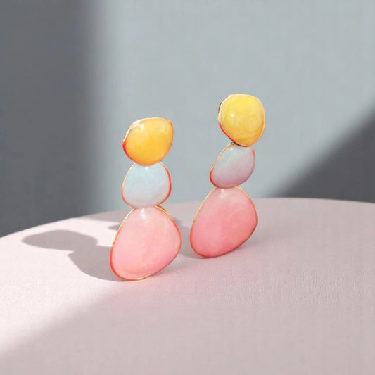 Colorful statement earrings featuring layered pastel stones in yellow, blue, and pink, displayed on a soft pink surface.