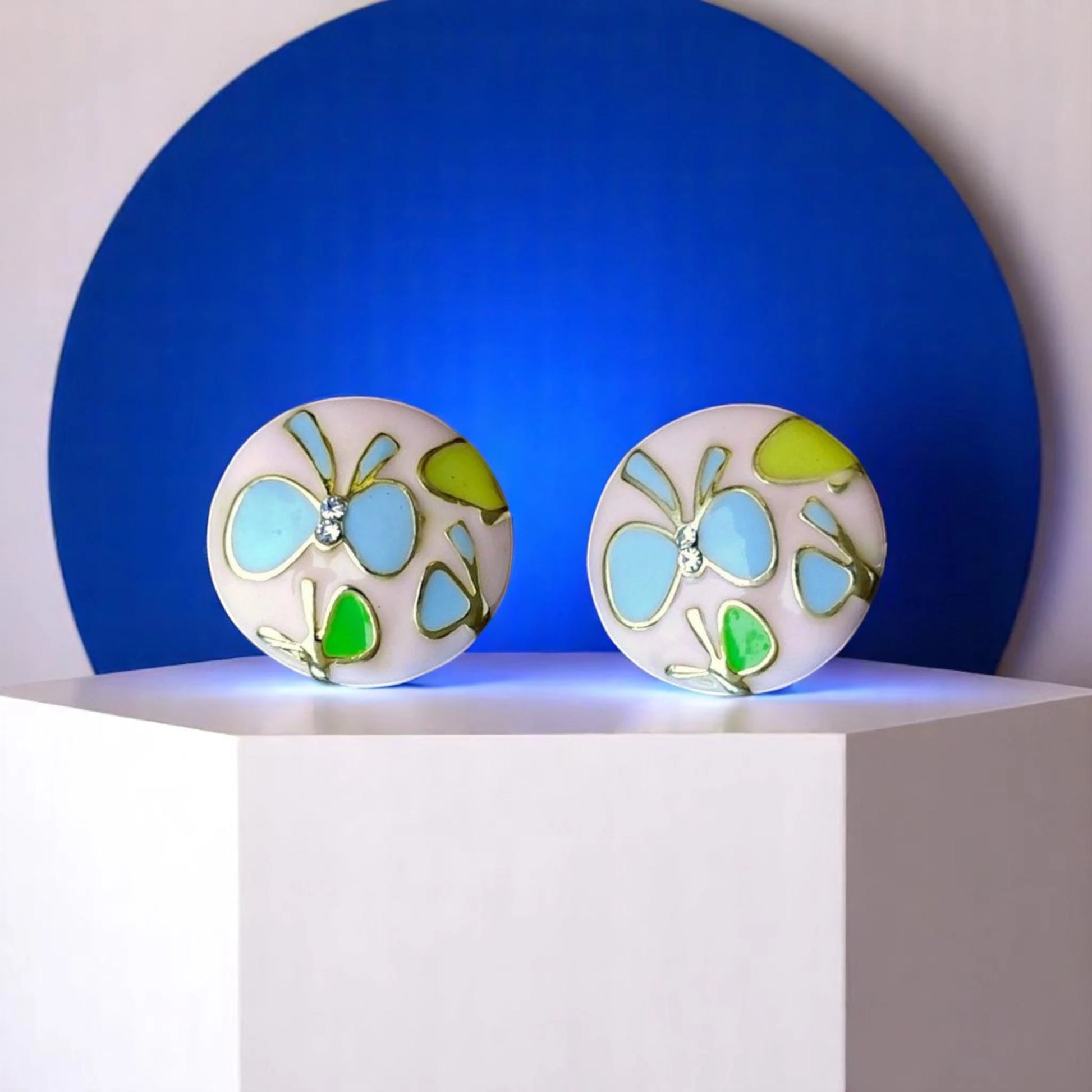 Two round decorative pieces with butterfly designs in blue and green, displayed on a pedestal against a blue background.