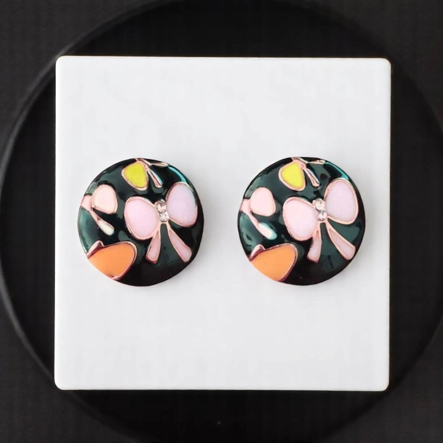 Colorful round earrings with floral designs, featuring pink, yellow, and orange on a dark background, displayed on a white platform.