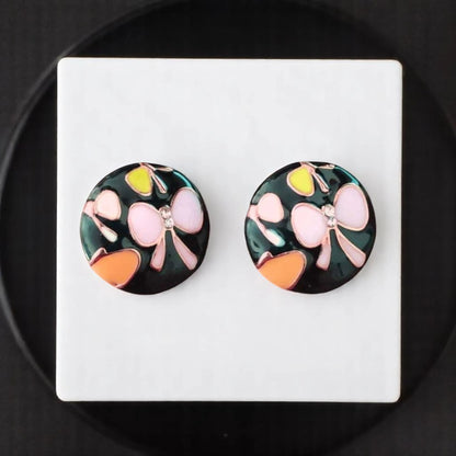 Colorful round earrings with floral designs, featuring pink, yellow, and orange on a dark background, displayed on a white platform.