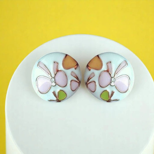 Colorful floral earrings with a glossy finish, featuring pastel blossoms and a tiny gem, displayed on a white stand against a yellow background.