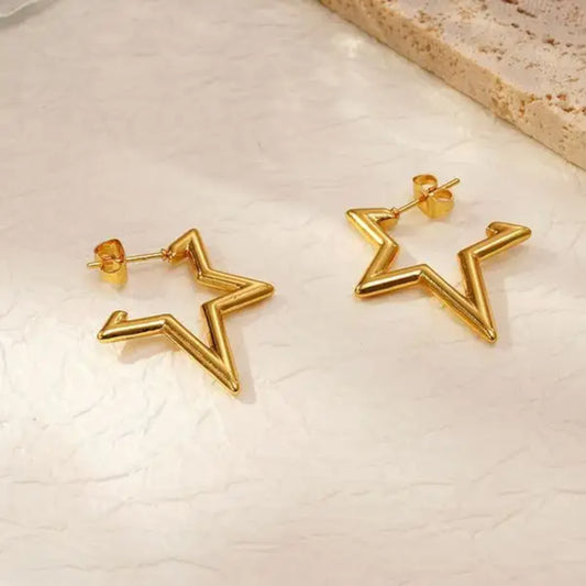 Gold star-shaped earrings with a sleek, angular design, resting on a textured surface.