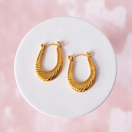 Golden spiral hoop earrings displayed on a white round pedestal, set against a soft pink background.