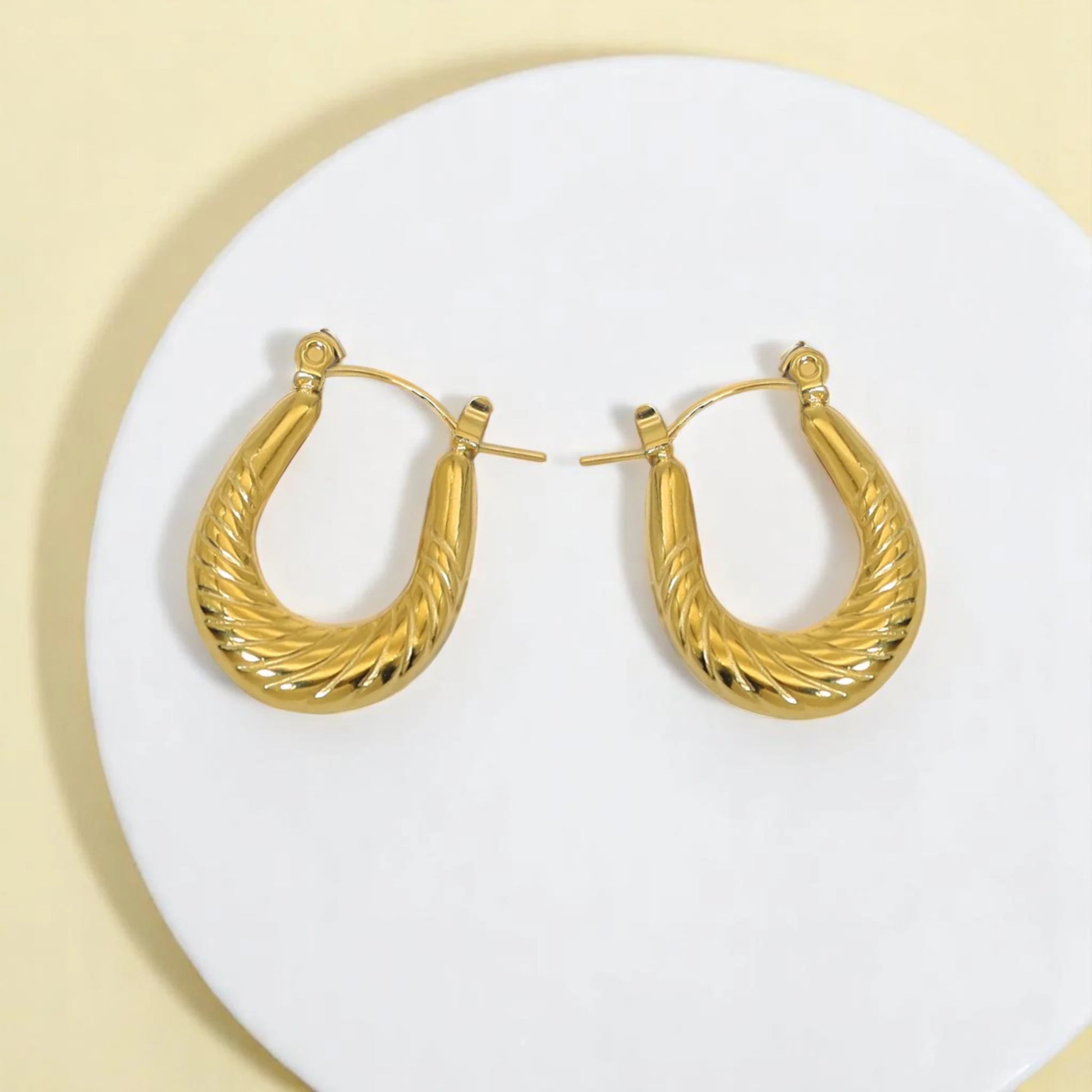 Shiny gold twisted hoop earrings displayed on a white round dish against a pale yellow background.