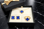A gold jewelry set with four clover-shaped motifs in deep blue enamel, displayed inside an open black gift box with a cream insert. The set includes a necklace, a pair of stud earrings, and a ring, with a matching black gift box and gold ribbon partially visible in the background on a black marble surface.