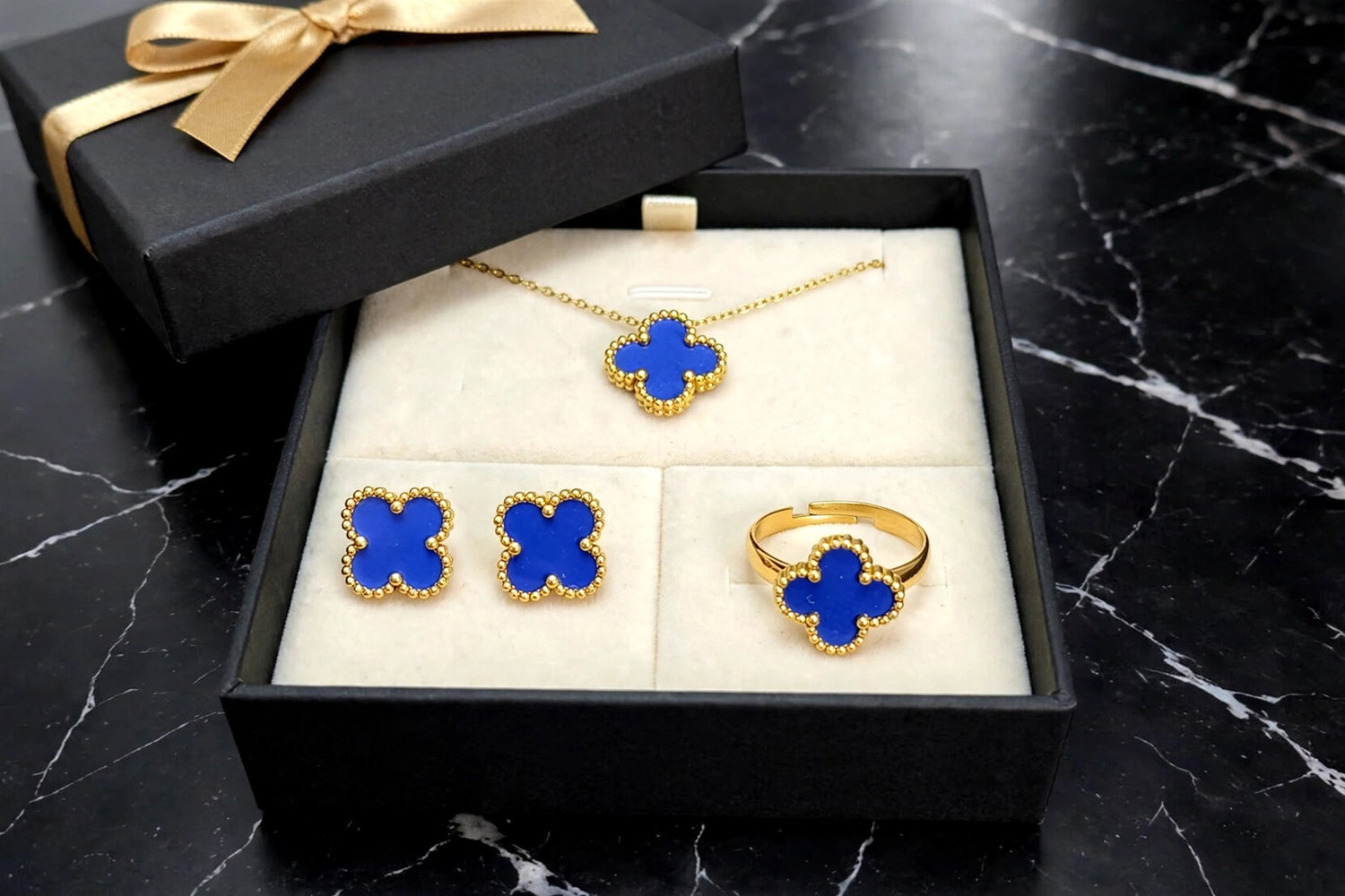 A gold jewelry set with four clover-shaped motifs in deep blue enamel, displayed inside an open black gift box with a cream insert. The set includes a necklace, a pair of stud earrings, and a ring, with a matching black gift box and gold ribbon partially visible in the background on a black marble surface.