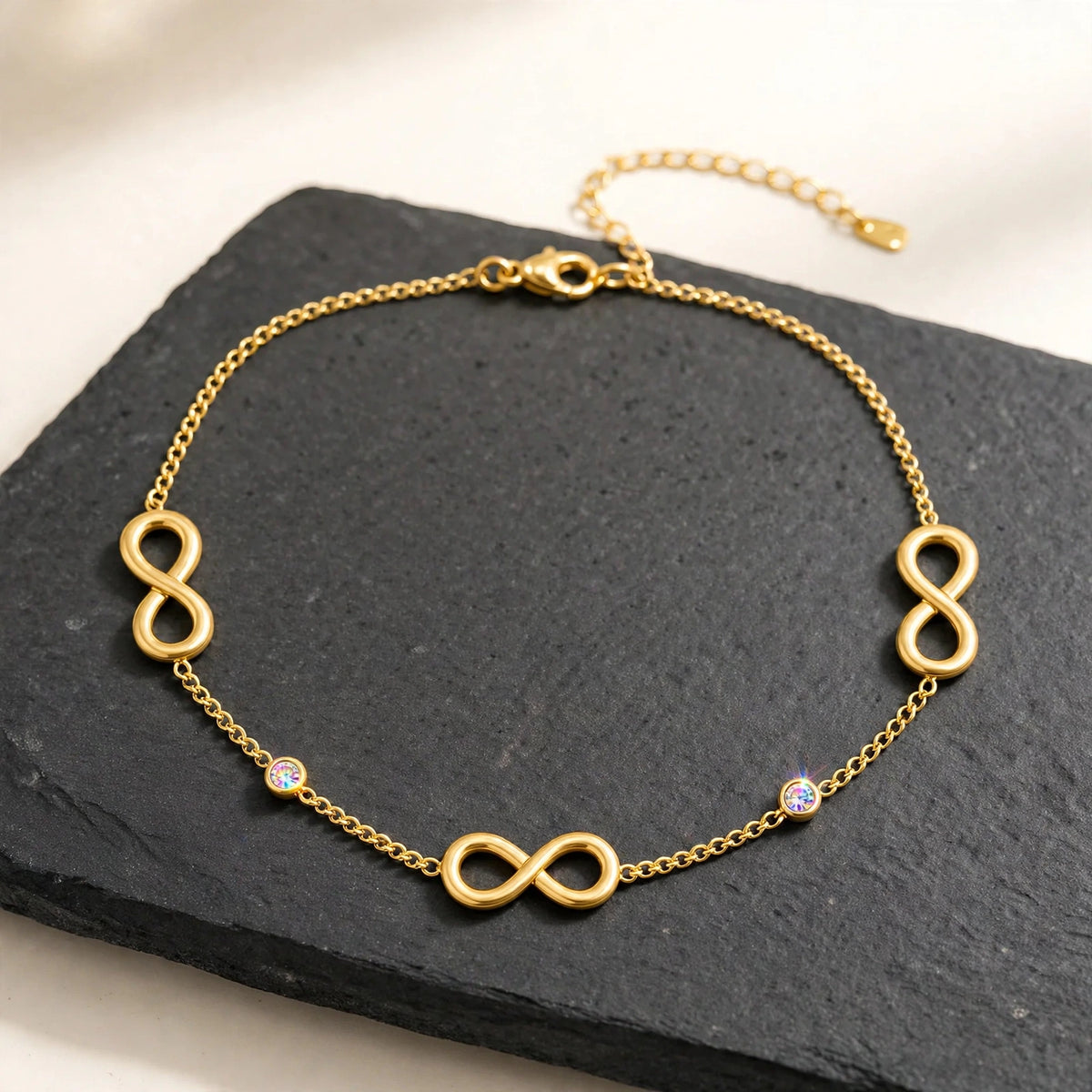 Gold infinity anklet with CZ stones on dark charcoal slate slab, high contrast.