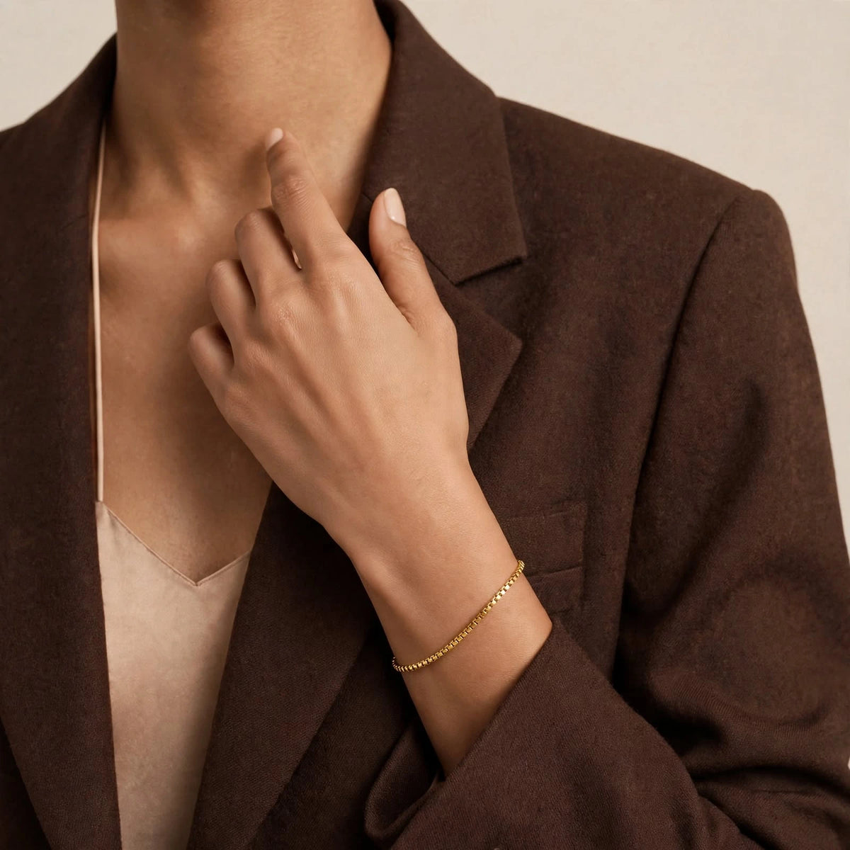 Woman in brown blazer touching collarbone with gold box chain bracelet on wrist