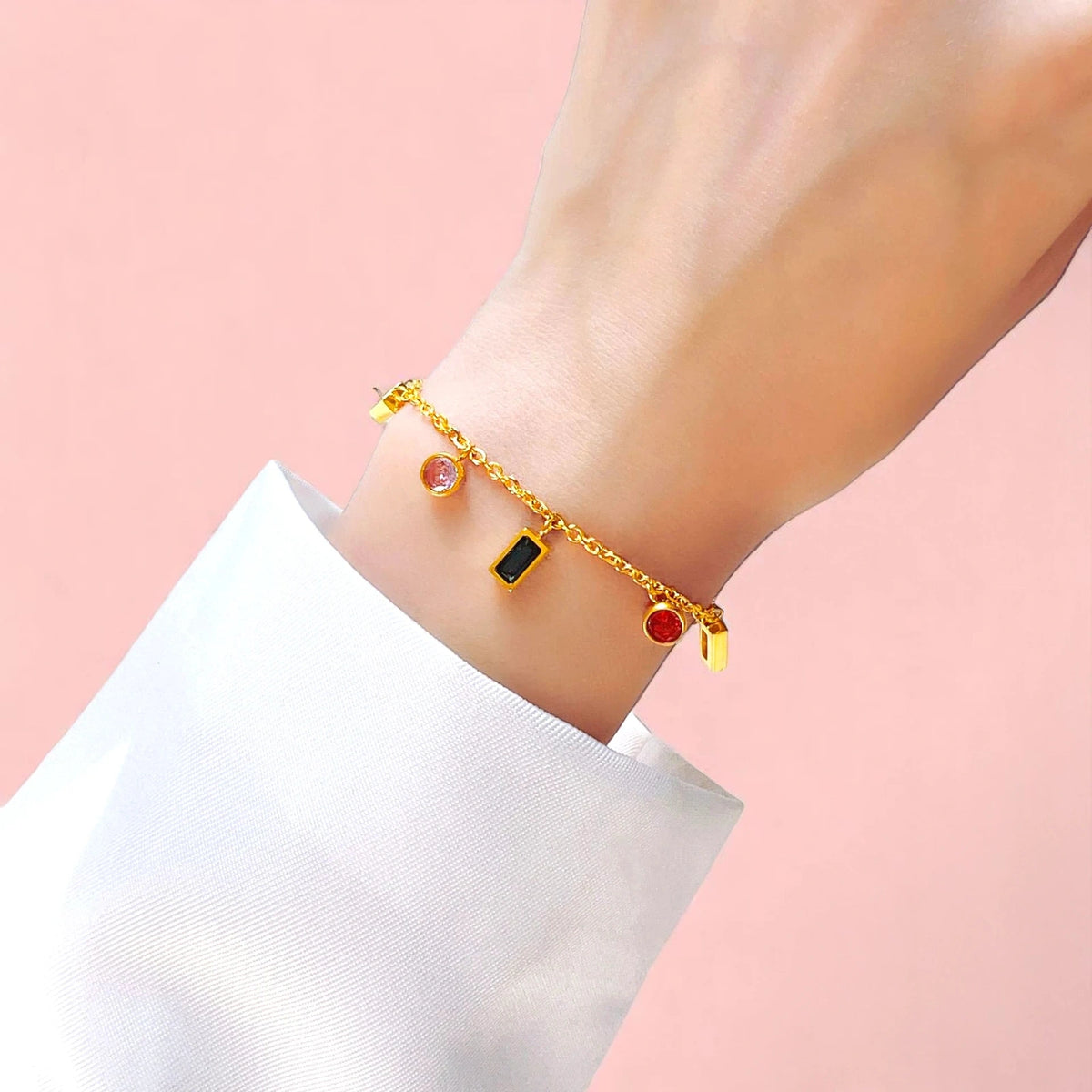 Gold charm bracelet with colorful round and baguette cubic zirconia charms on wrist, white sleeve.