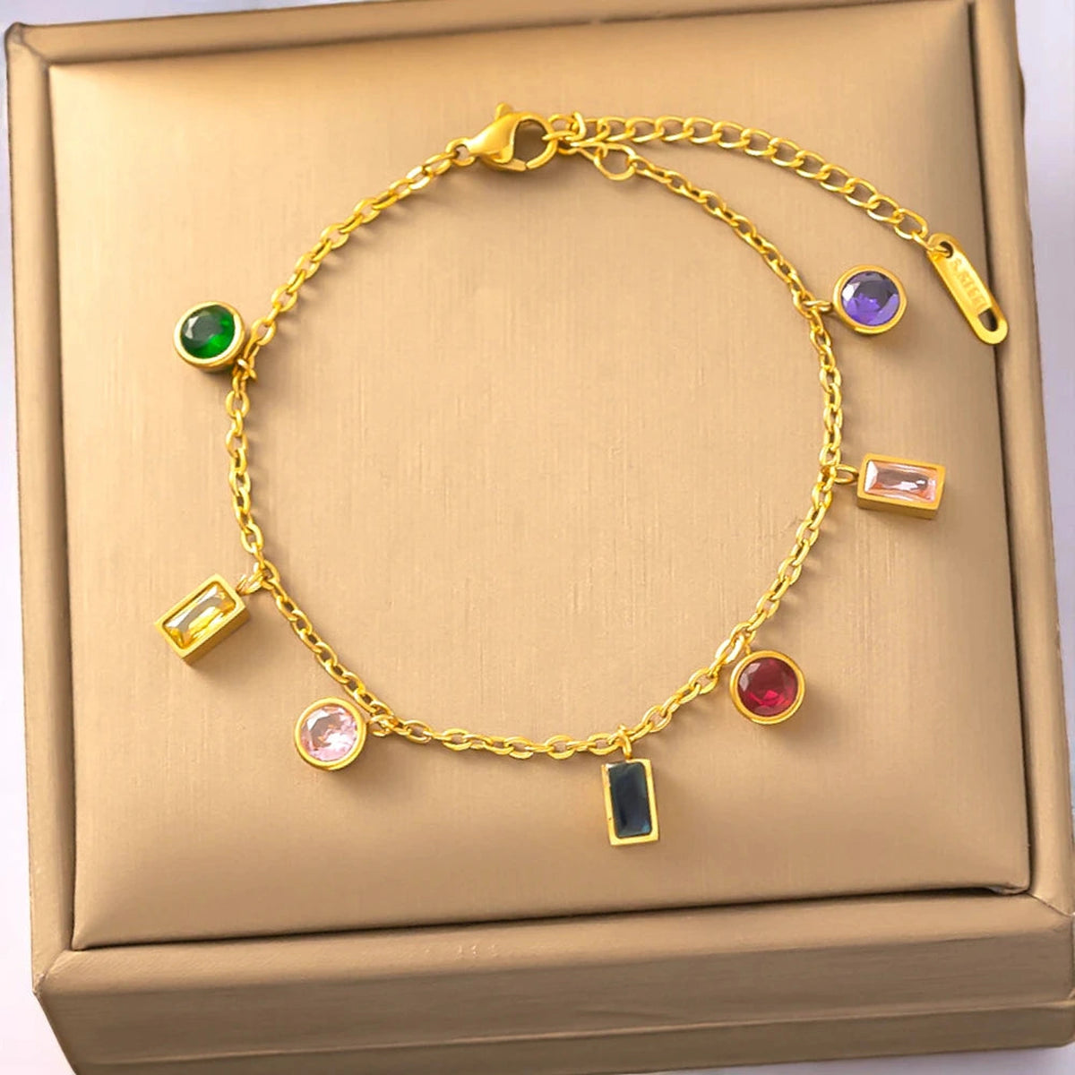 Gold multicolor gemstone charm bracelet on beige velvet tray against warm white background.