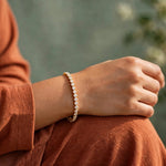 Woman's wrist with gold tennis bracelet resting on knee, terracotta top, sage green backdrop