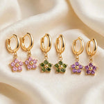 Three pairs of Jespara gold huggie hoop earrings with flower charms in pink, olive green and purple crystals on cream silk background