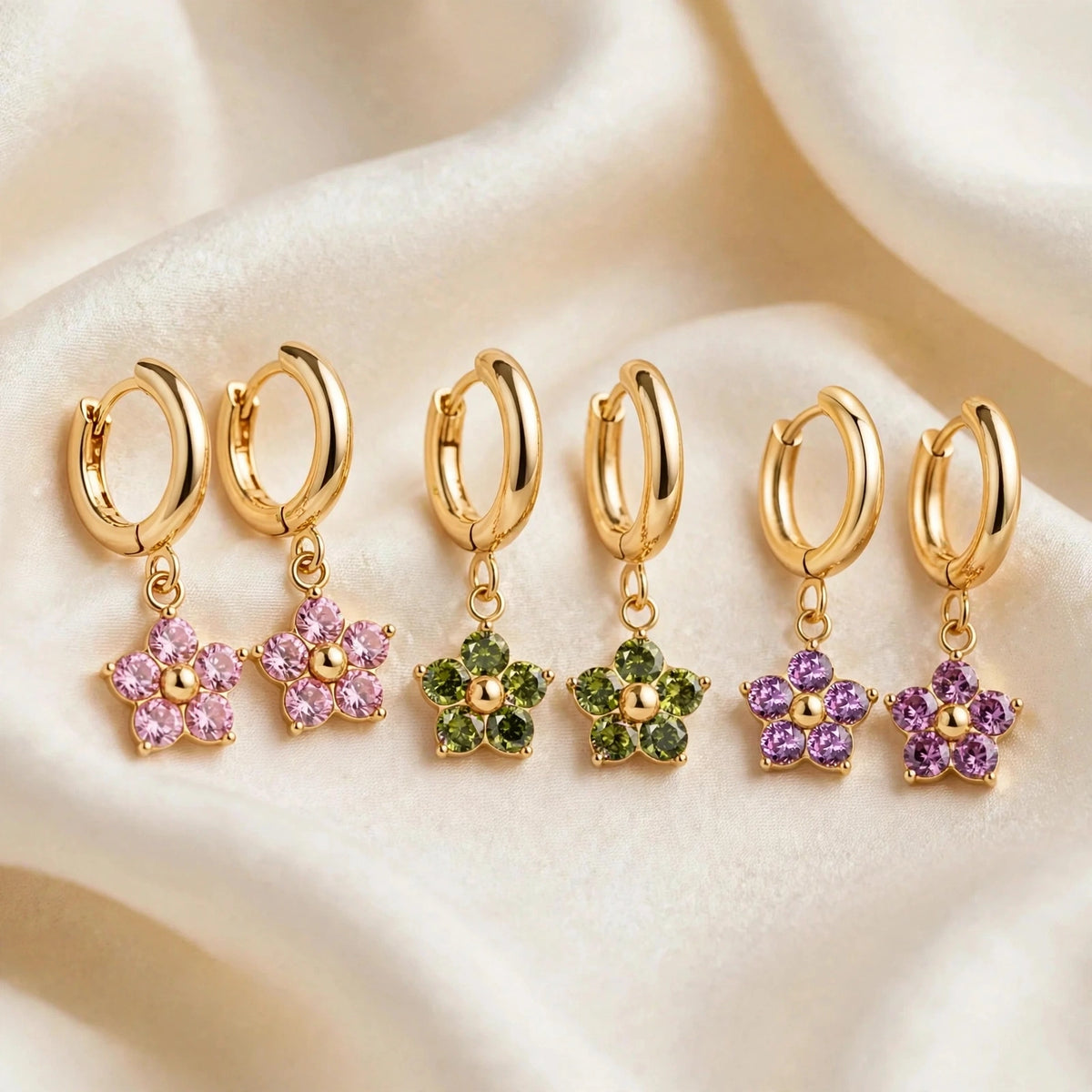 Three pairs of Jespara gold huggie hoop earrings with flower charms in pink, olive green and purple crystals on cream silk background