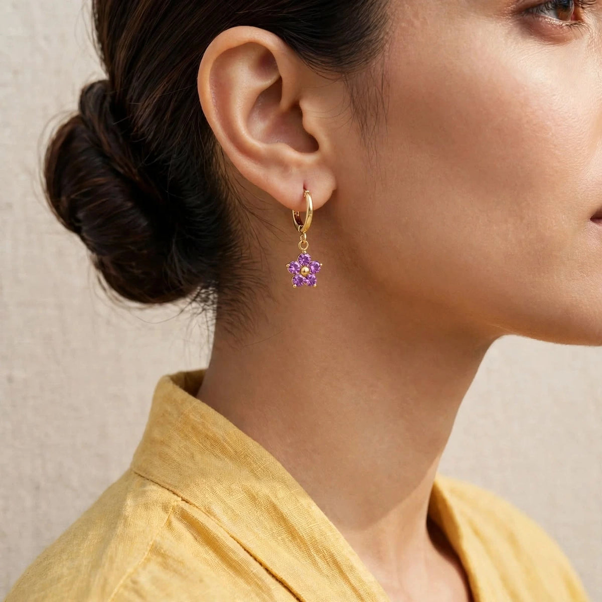 Smaller purple flower huggie earring on ear, yellow wrap blouse, low bun