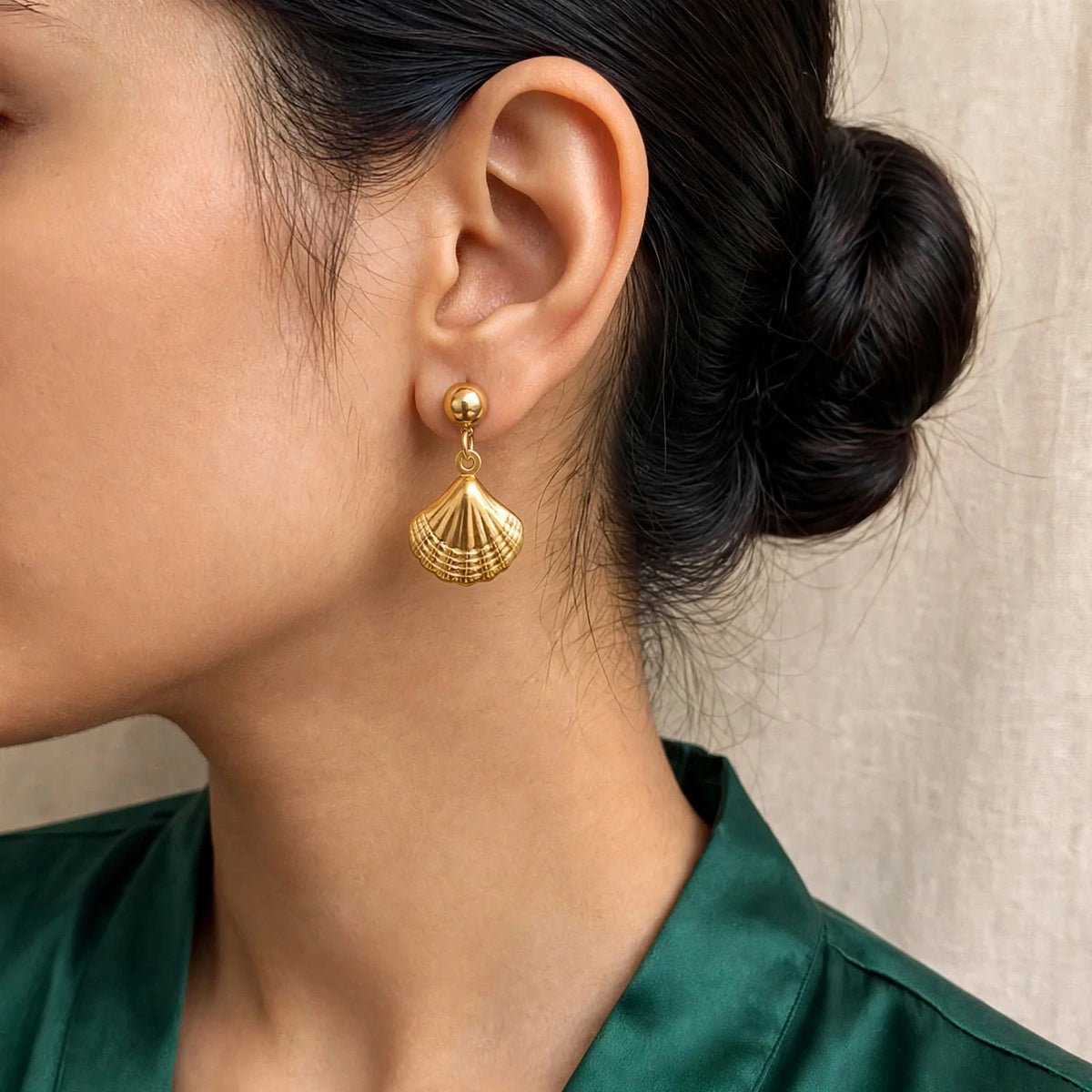 Gold shell drop earring on model with low bun and emerald blouse, close-up ear view.