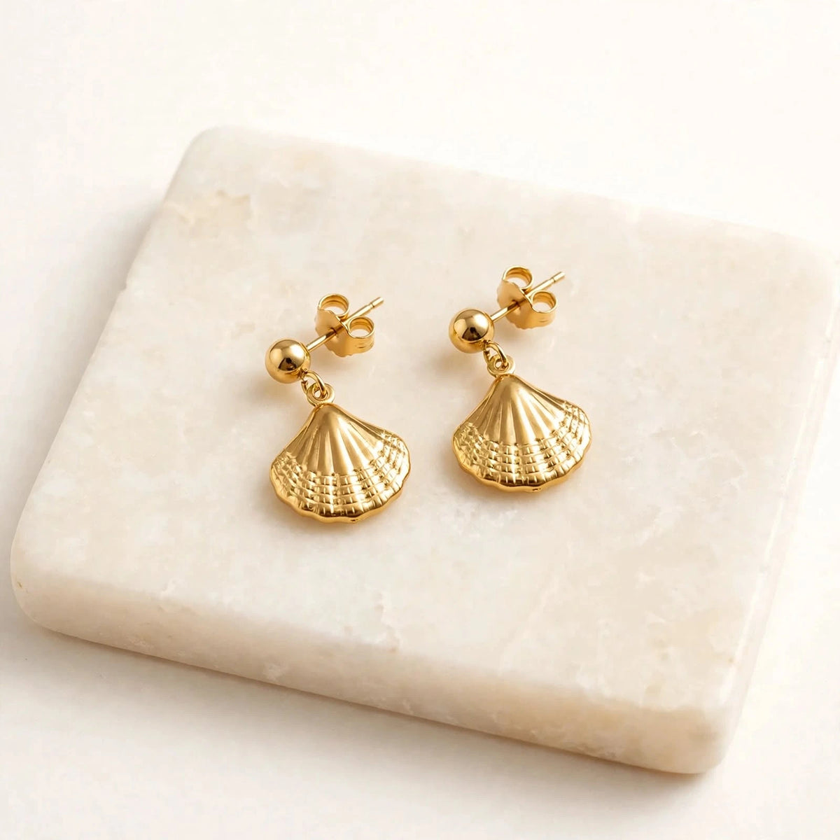 Gold shell drop earrings pair on ivory stone plinth, warm-white background.
