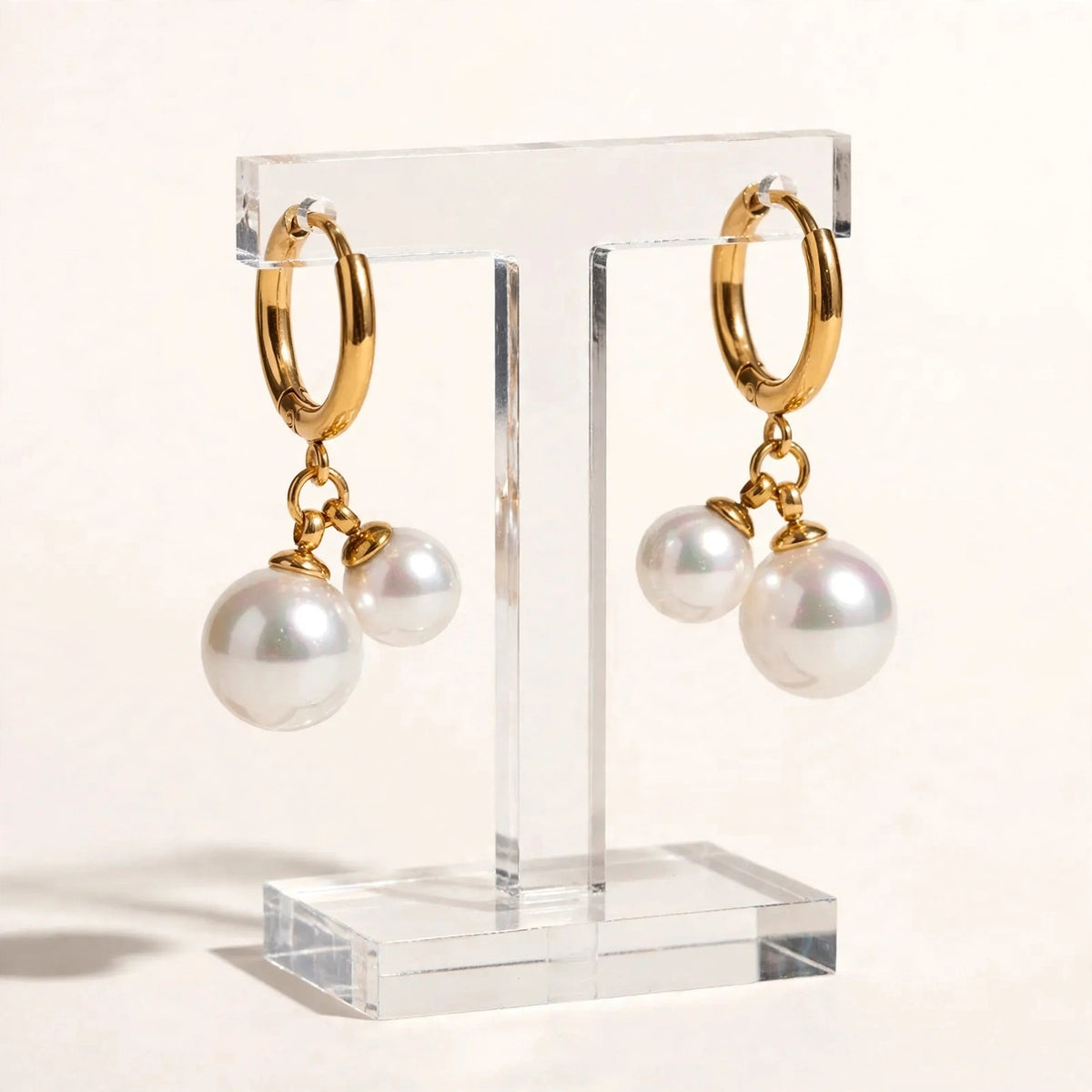 Gold huggie hoop double pearl earrings on clear acrylic stand, warm white backdrop