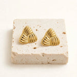 Gold triangular ribbed earrings pair on cream travertine block on warm-white background.