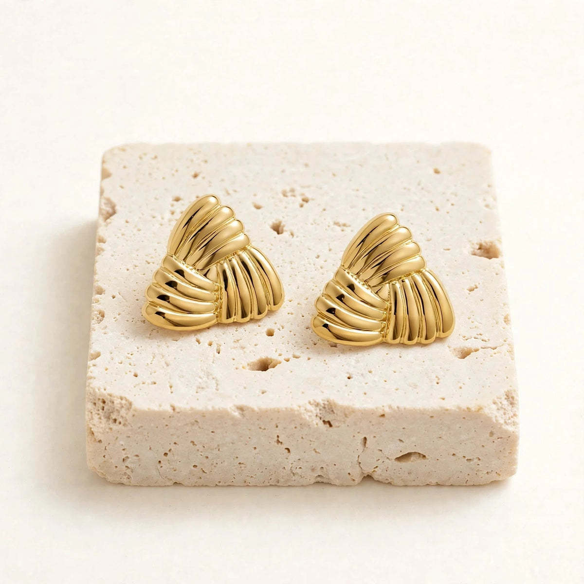 Gold triangular ribbed earrings pair on cream travertine block on warm-white background.