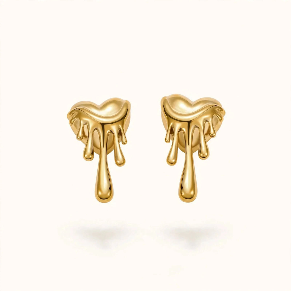 Gold melting heart drip earrings pair floating on warm off-white background.