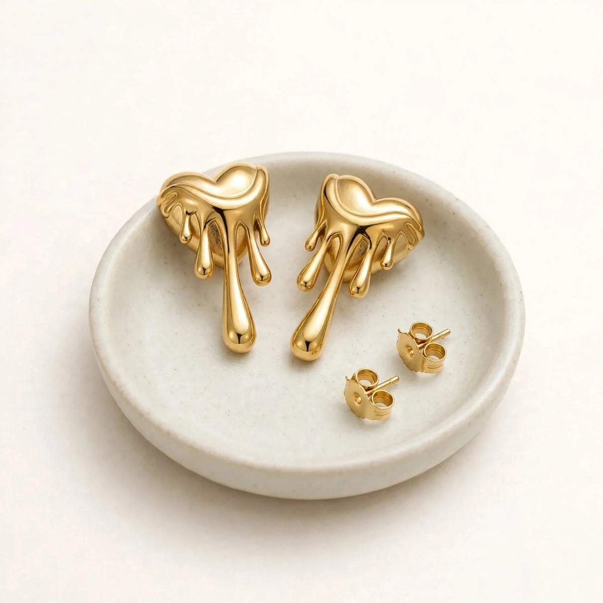 Gold melting heart drip earrings pair on small pale ceramic dish on warm-white background.