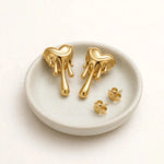 Gold melting heart drip earrings pair on small pale ceramic dish on warm-white background.
