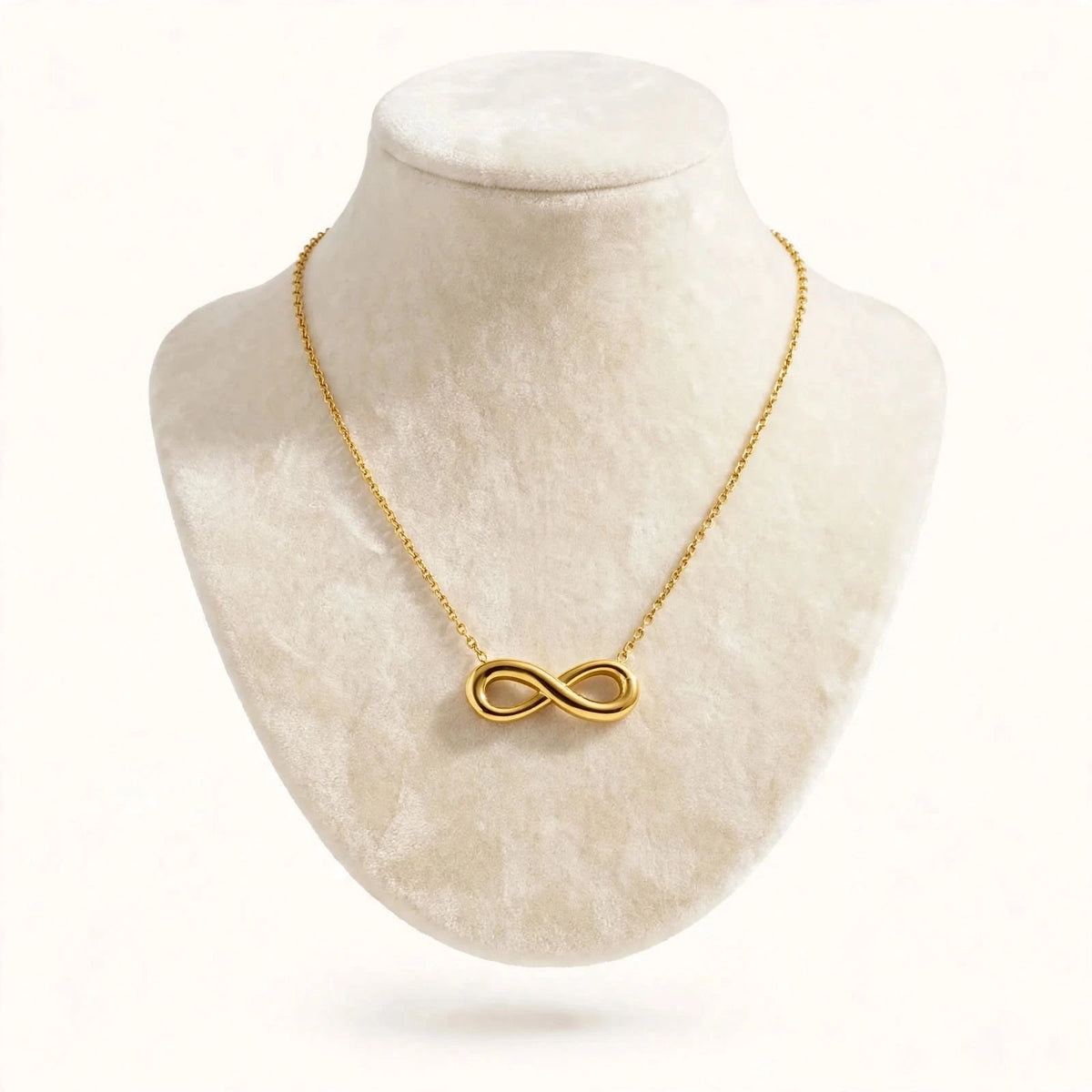 Gold infinity necklace on ivory velvet bust against warm-white background