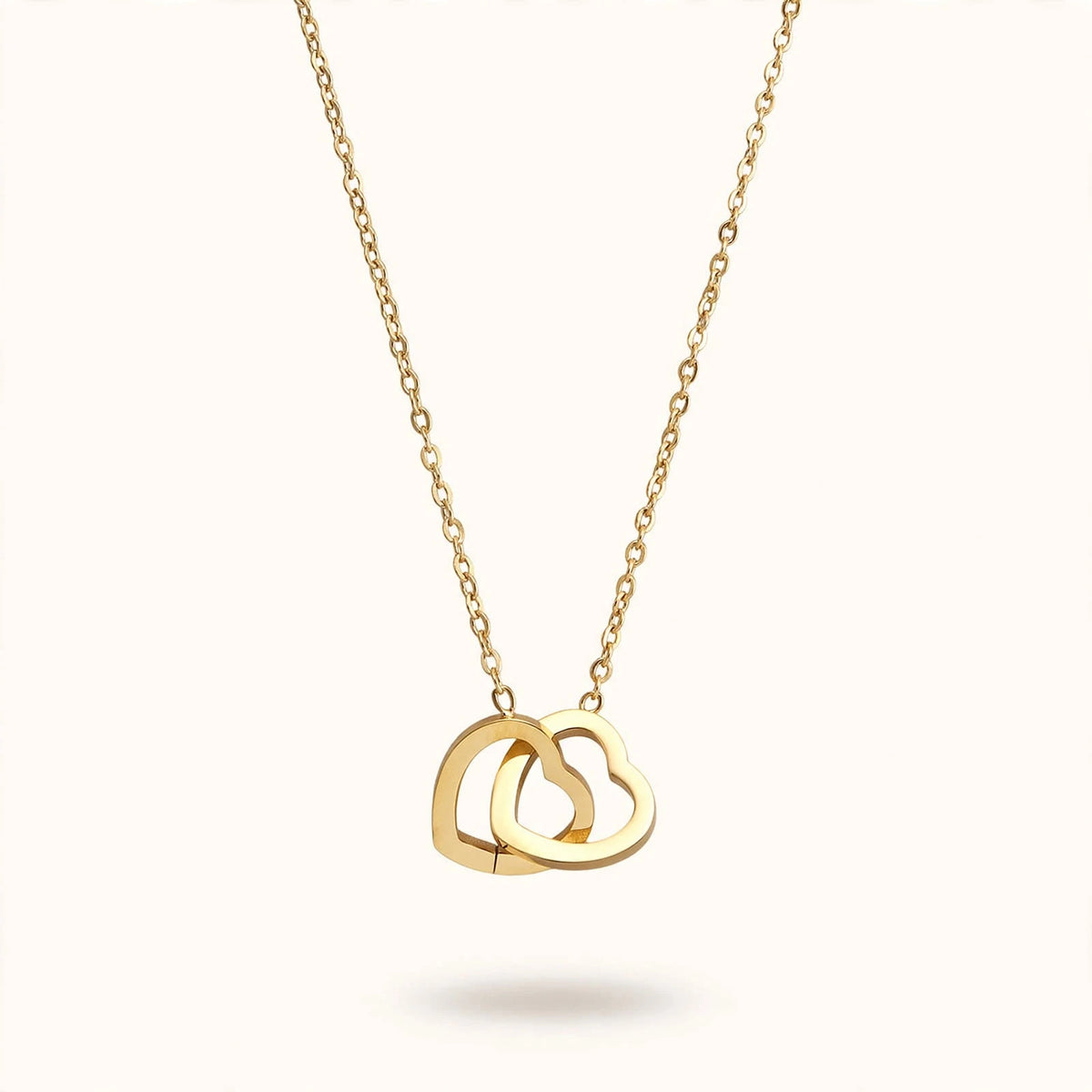Gold double interlocked heart pendant necklace floating on warm off-white background.