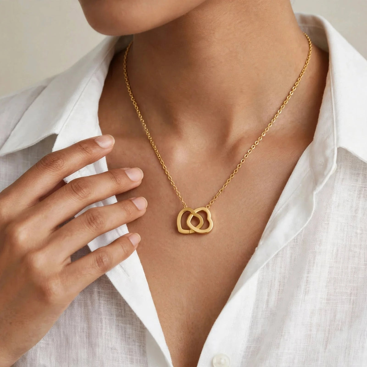 Indian woman in white linen shirt wearing gold double heart pendant, hand near collarbone.