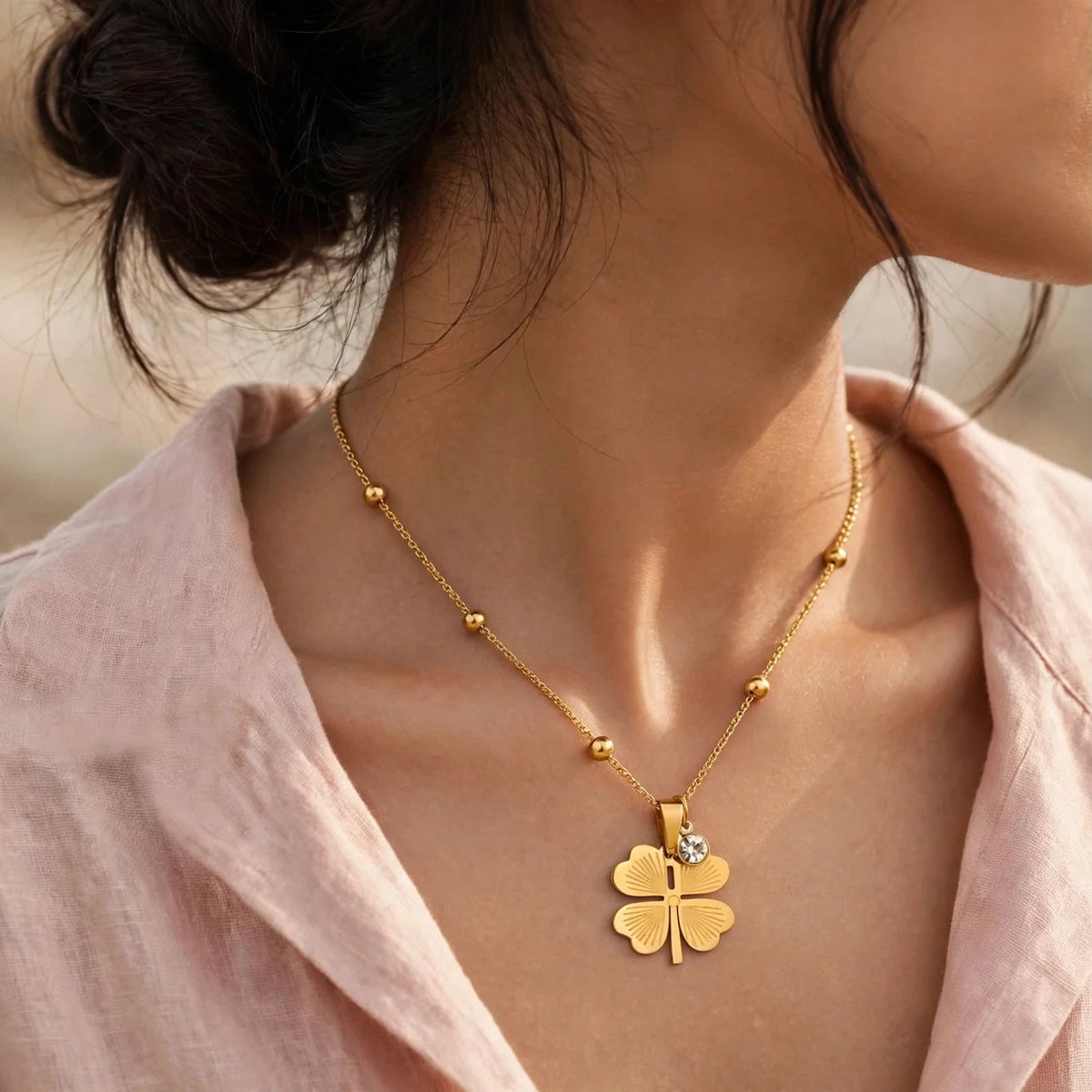 Woman in soft blush wrap blouse wearing gold four-leaf clover necklace turned slightly