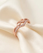 Rose gold infinity ring with CZ stones resting on ivory silk against a warm-white background.