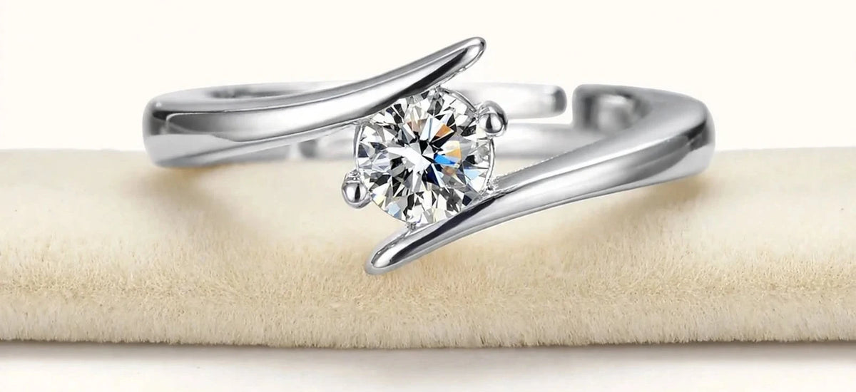 Silver open bypass solitaire ring lying horizontal on slim cream velvet cushion.