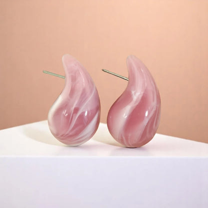 Two pink marbled teardrop earrings displayed on a white pedestal, set against a soft peach background.