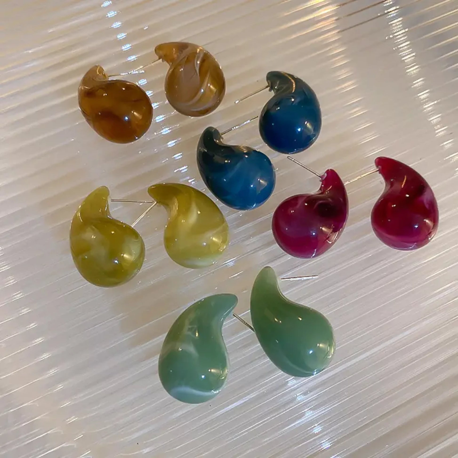 A collection of colorful, teardrop-shaped earrings displayed on a textured surface, featuring shades of brown, blue, green, yellow, and pink.