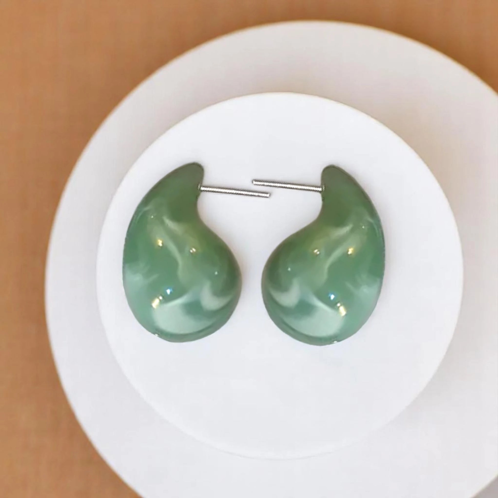 Green marbled drop earrings displayed on a white circular dish against a warm background. 