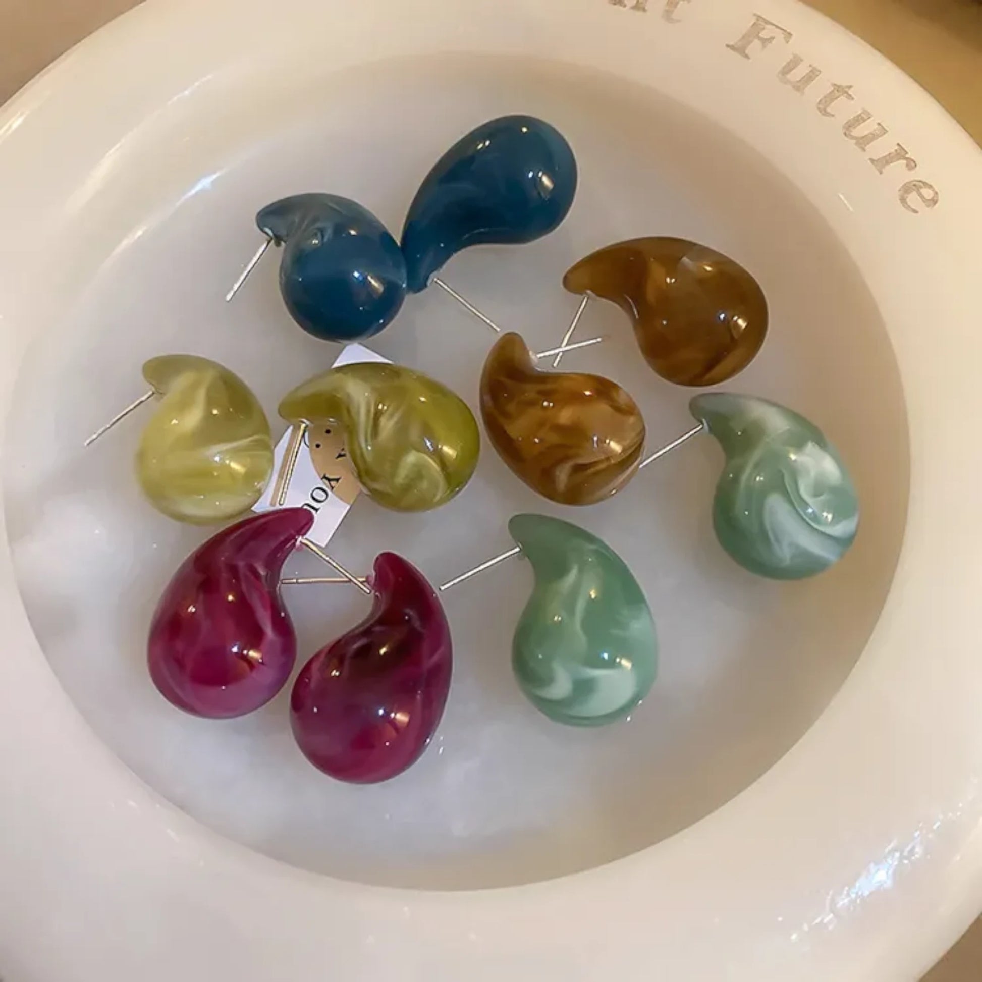 A collection of colorful, teardrop-shaped earrings displayed on a textured surface, featuring shades of brown, blue, green, yellow, and pink.