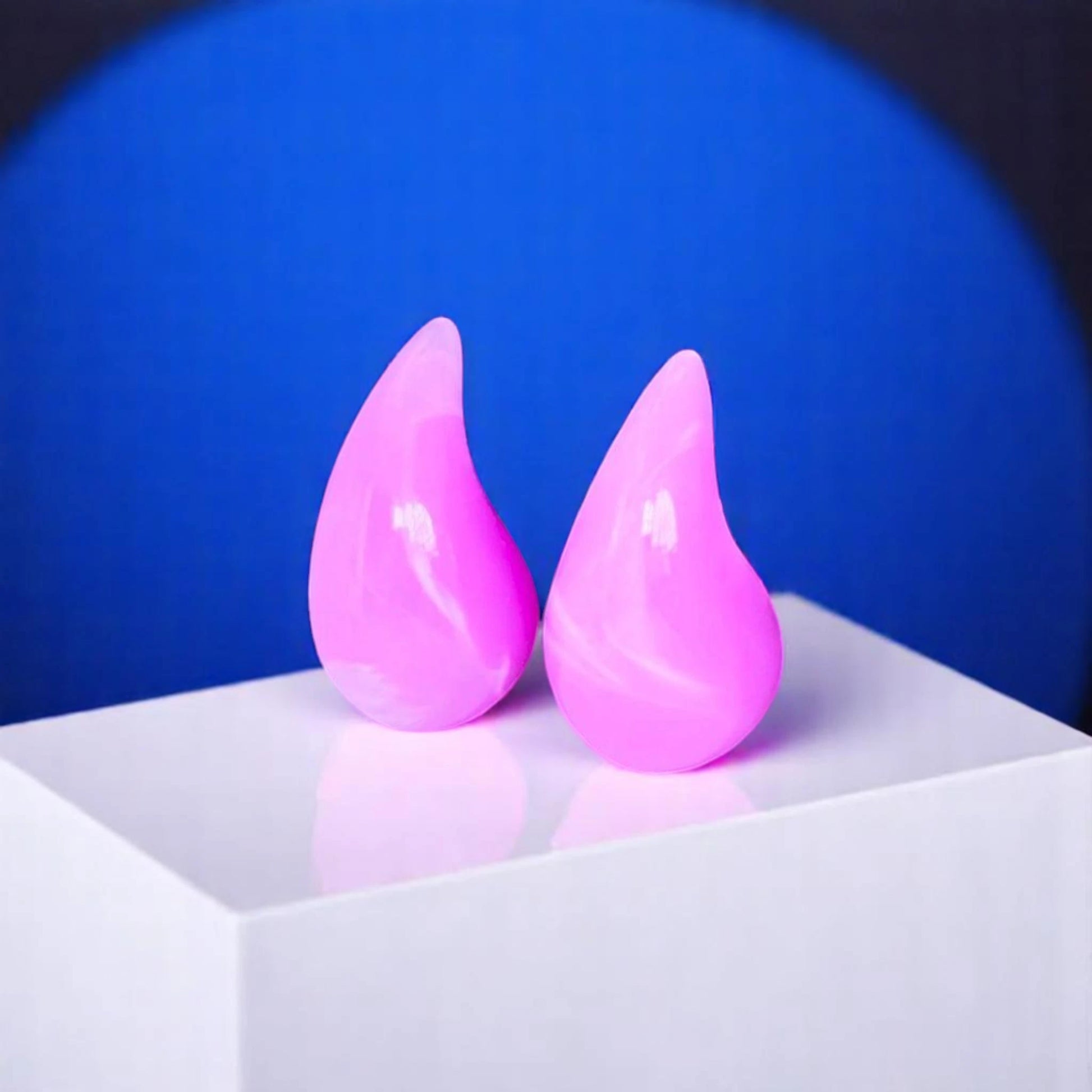 Two glossy, pink teardrop-shaped objects sit on a white pedestal against a blue background, reflecting light beautifully.
