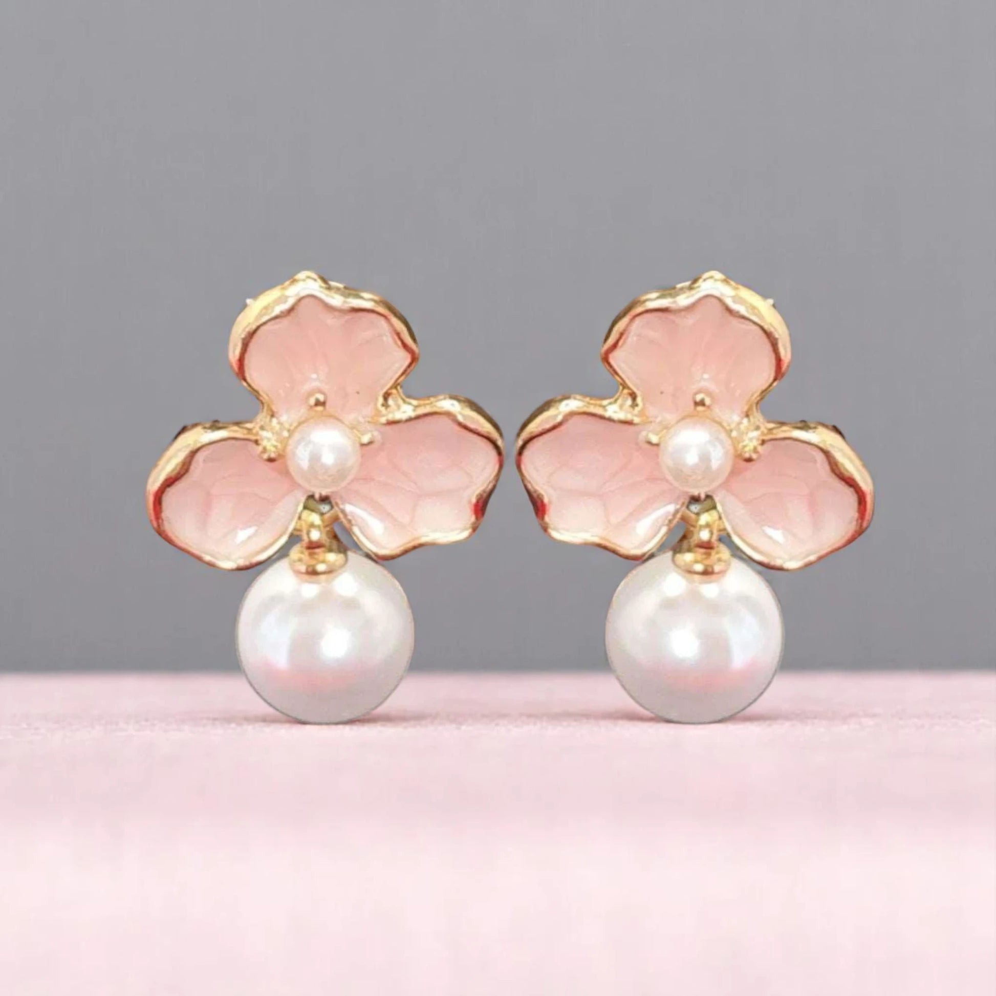 Delicate earrings featuring pink floral designs with gold trim and a central white pearl, elegantly displayed on a soft pink background.