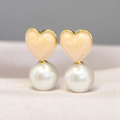 Gold heart-shaped earrings topped with peach enamel, featuring a lustrous white pearl drop. Elegant and playful design.