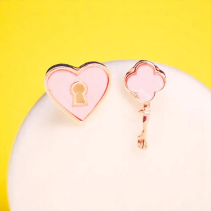Pink heart-shaped and key-shaped earrings with gold accents, displayed on a white stand against a vibrant yellow background.