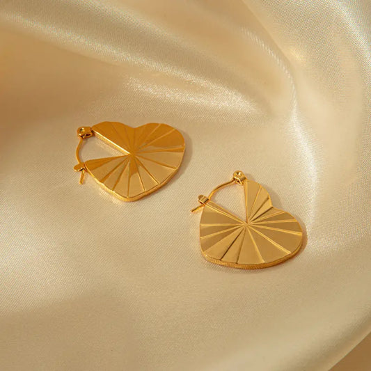 Two golden heart-shaped earrings with a geometric pattern resting on a smooth, light-colored satin fabric.