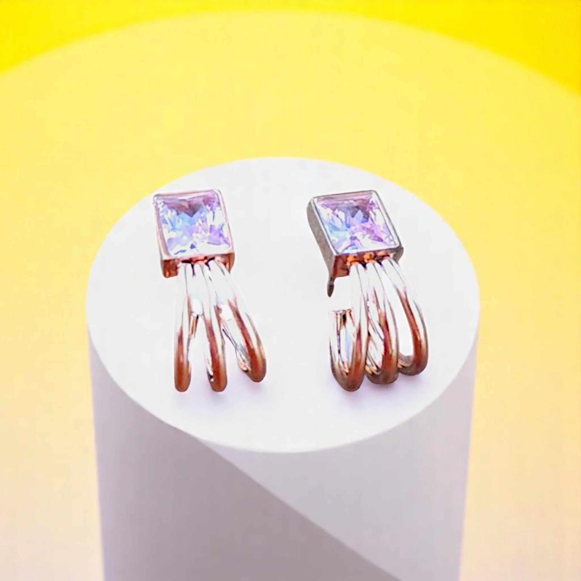A pair of elegant earrings featuring square purple gemstones and intertwined silver and rose gold strands, displayed on a pedestal.
