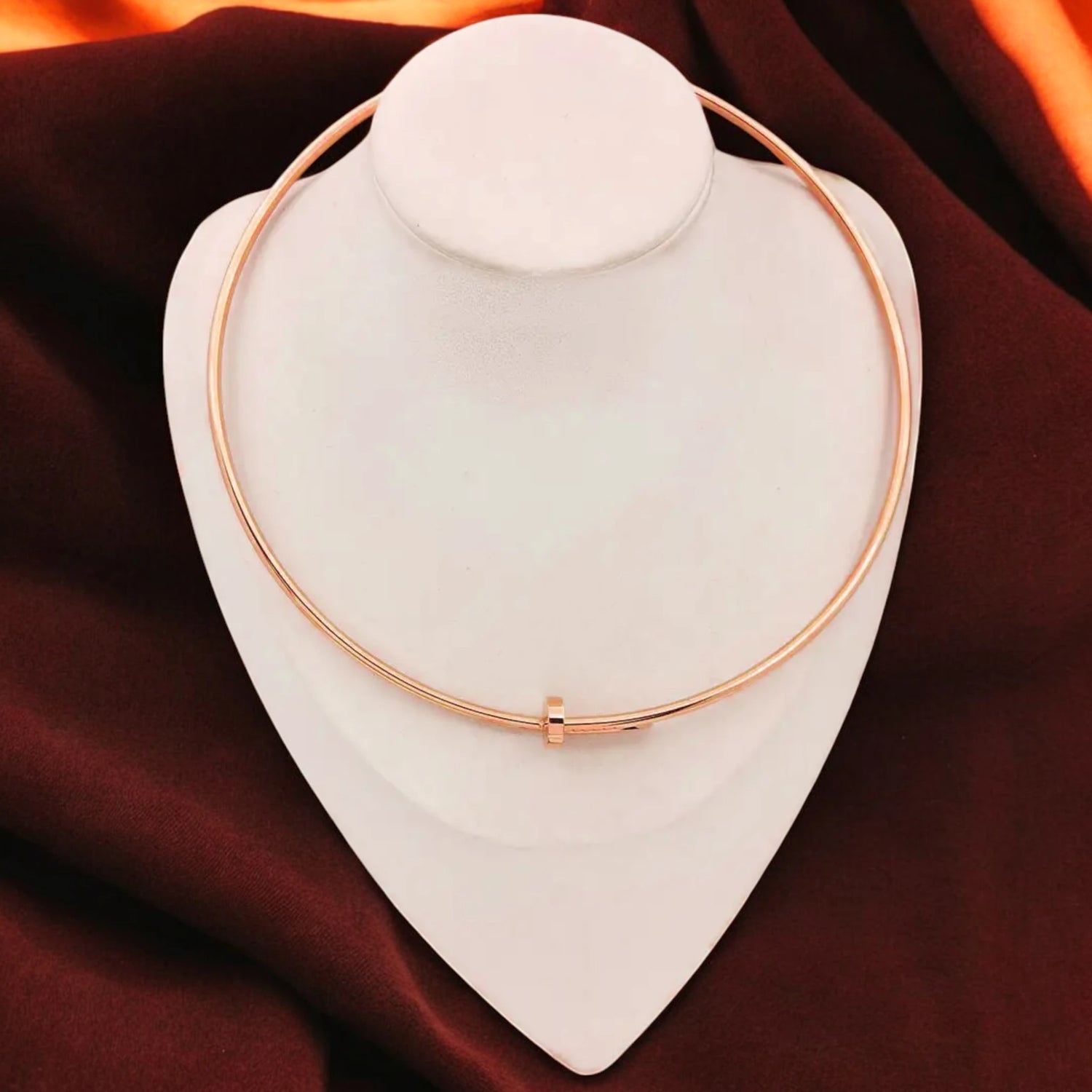 An elegant rose gold nail choker necklace is displayed on a white jewelry stand against a rich maroon and orange fabric background, highlighting minimalist and modern design.
