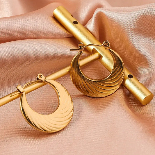 Golden earrings with a wavy design resting on a golden rod atop a soft pink satin background.