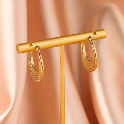 A pair of elegant gold hoop earrings hanging on a gold display stand against a soft, blush pink satin background.